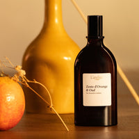 Black bottle of 'Zeste d'Orange & Oud' by L'Atelier on a wooden surface with an orange and vase in the background.